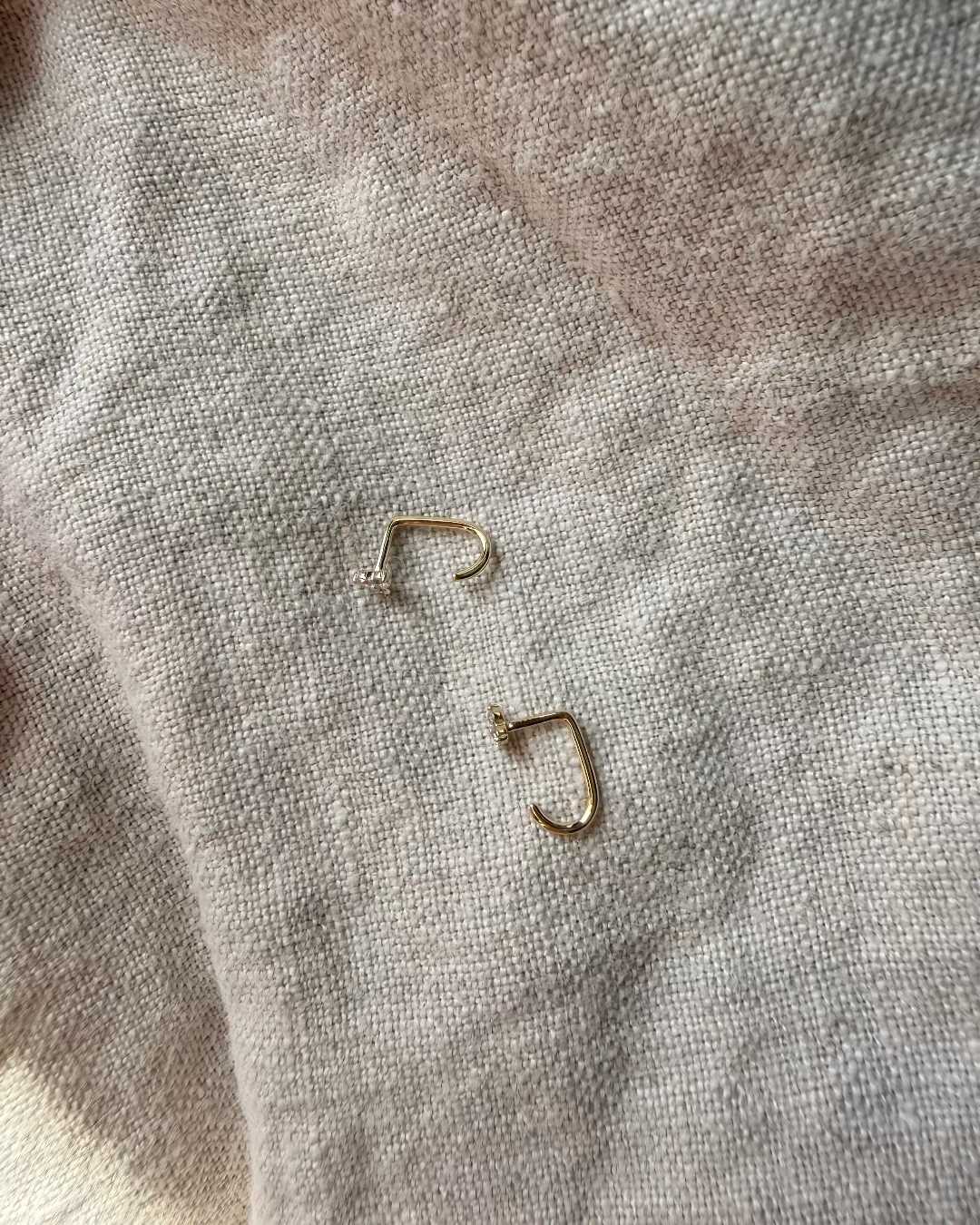 Two gold earrings on a textured beige fabric surface